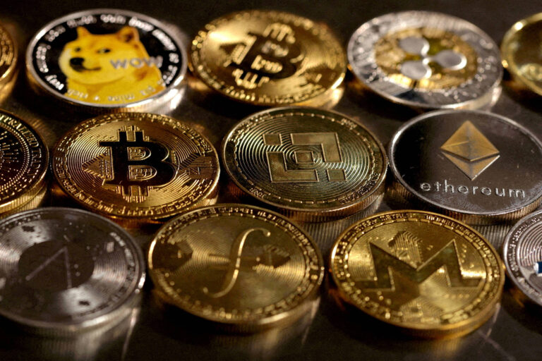 A imagem mostra uma variedade de moedas de criptomoedas dispostas em uma superfície. Entre elas, estão moedas com os logotipos de Bitcoin, Ethereum, Dogecoin, e outras criptomoedas. As moedas têm acabamentos metálicos, algumas em ouro e outras em prata, refletindo a luz. A disposição é organizada, com algumas moedas visíveis em diferentes ângulos.