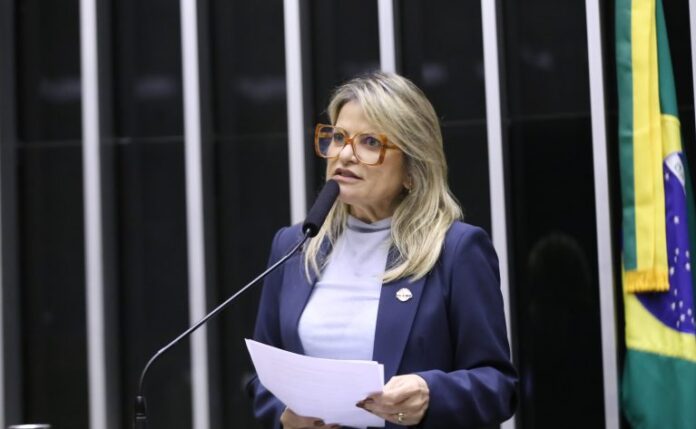 Deputada Flávia Morais fala ao microfone. Ela é loira, tem o cabelo comprido, usa uma blusa branca e segura um papel