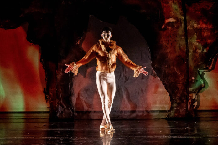 Bailarino de pé no centro do palco com braços abertos, usando casaco marrom e calça branca justa. Fundo escuro com iluminação em tons quentes destaca a figura.