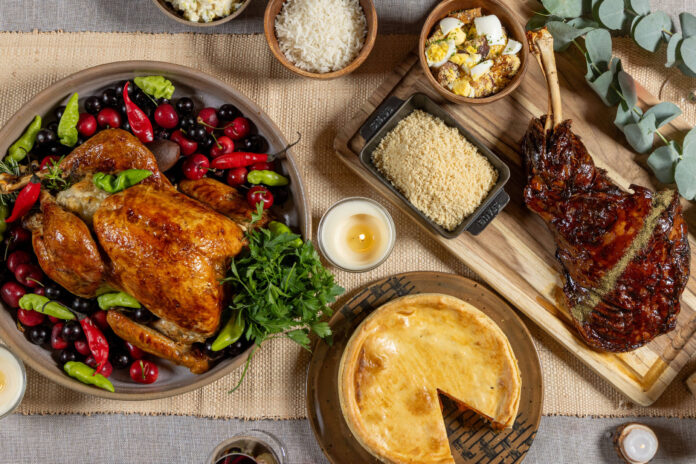Mesa posta com peru assado decorado com frutas vermelhas e pimentas, pernil assado sobre tábua de madeira, torta com uma fatia retirada, e tigelas com arroz, pipoca e outros acompanhamentos.