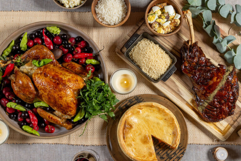 Mesa posta com peru assado decorado com frutas vermelhas e pimentas, pernil assado sobre tábua de madeira, torta com uma fatia retirada, e tigelas com arroz, pipoca e outros acompanhamentos.