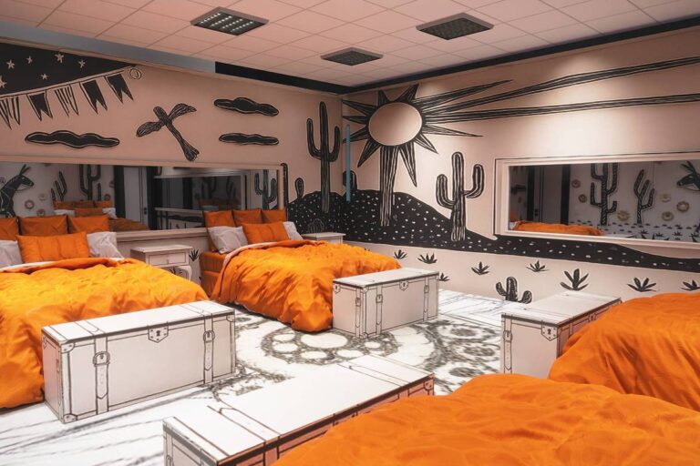 Quarto com três camas de casal com edredons laranja vibrante. Paredes e chão decorados com desenhos em preto que simulam cactos, sol, estrelas e elementos do deserto. Três baús brancos com contornos pretos ficam ao pé das camas. Espelhos grandes refletem a decoração temática.