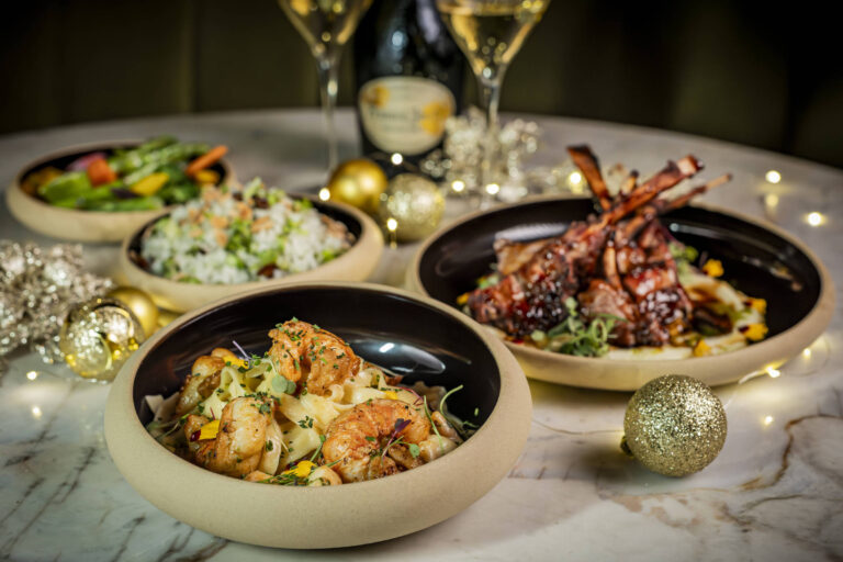 Três pratos gourmet servidos em tigelas claras com interior preto sobre mesa de mármore. À frente, prato com camarões grelhados sobre risoto cremoso. Ao fundo à direita, costeletas de cordeiro com molho escuro e guarnição. Ao fundo à esquerda, salada colorida com vegetais variados. Garrafa de espumante e duas taças ao centro, com decoração natalina dourada ao redor.