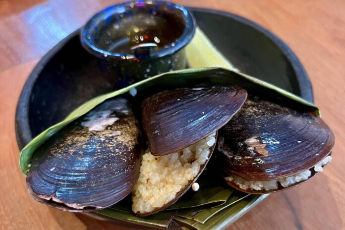 Três mariscos escuros servidos em folha verde dobrada, acompanhados de pequeno recipiente com molho escuro, sobre prato preto em mesa de madeira.