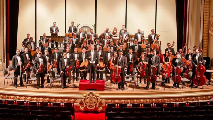 Orquestra sinfônica reunida no palco de um teatro histórico, com músicos e maestro em formação clássica, simbolizando a força da música como expressão do Espírito do Natal e da comunhão humana.