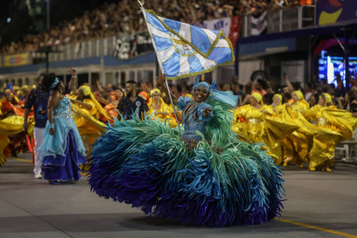 Uma pessoa vestida com um traje colorido e volumoso, predominantemente azul e verde, dança enquanto segura uma bandeira. Ao fundo, há um grupo de pessoas vestindo trajes amarelos e outras cores, todos participando de um desfile. A cena é animada, com uma grande multidão assistindo ao evento.
