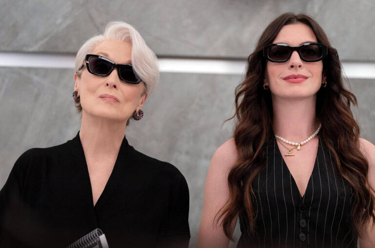 Duas mulheres posam lado a lado contra fundo cinza. A da esquerda tem cabelo branco curto, veste preto e usa brincos grandes. A da direita tem cabelo longo castanho, veste colete listrado e colares de pérolas.
