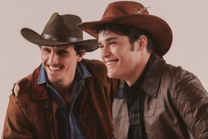 Dois homens sentados lado a lado, ambos usando chapéus de cowboy e jaquetas marrons. Eles sorriem olhando para a direita, com fundo neutro.