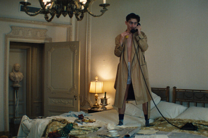 Homem de sobretudo bege está em pé sobre cama desarrumada em quarto com decoração clássica. Ele segura um telefone antigo com fio, ligado a um suporte no teto. O quarto tem paredes claras, duas luminárias de mesa acesas e uma porta aberta que revela uma estátua em um corredor.