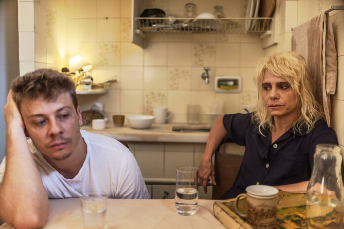 Homem e mulher sentados à mesa de cozinha pequena, ambos com expressão séria e distante. Mesa tem copos de água, xícara e jarra. Ambiente simples com azulejos brancos e utensílios ao fundo.