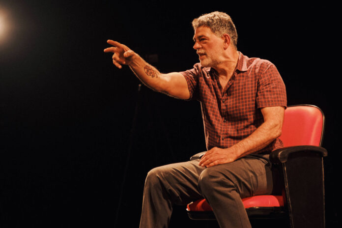 Homem de meia-idade sentado em cadeira vermelha em palco escuro, vestindo camisa xadrez vermelha e calça cinza, apontando com o braço direito estendido para frente.