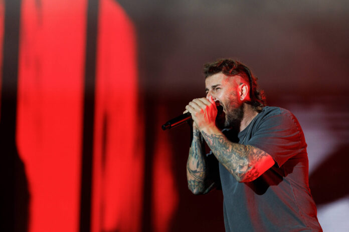 Avenged Sevenfold faz show em São Paulo - 28/01/2026 - Homem com barba e tatuagens nos braços segura microfone próximo à boca enquanto canta em palco com fundo iluminado por luz vermelha intensa.