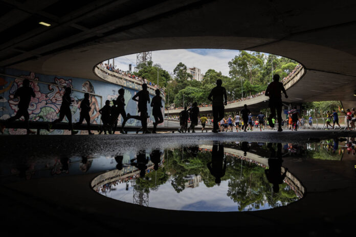 Diversas pessoas caminham sob um viaduto com abertura circular, onde a luz natural entra. No chão, uma poça d'água reflete as silhuetas das pessoas e parte da estrutura do viaduto. Ao fundo, há árvores, edifícios e um muro com grafite colorido.