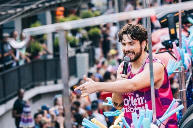 Homem sorridente com camiseta rosa distribui brindes para multidão durante desfile em rua movimentada. Pessoas observam e interagem ao redor, com decoração colorida e elementos festivos visíveis.
