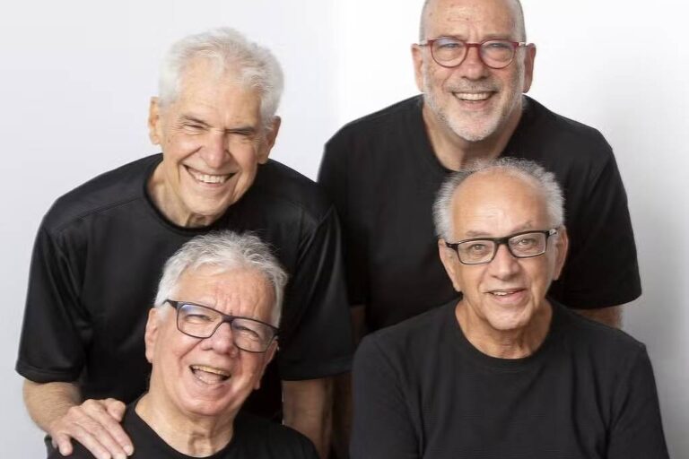 Quatro homens idosos vestindo camisetas pretas posam juntos contra fundo branco. Dois estão sentados à frente e dois em pé atrás, todos usando óculos e sorrindo.