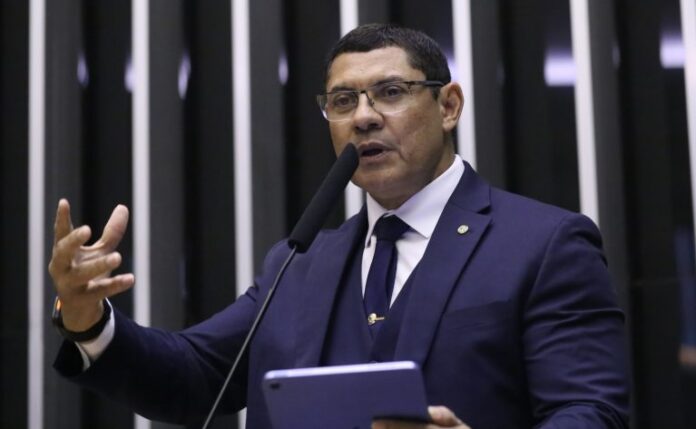 Deputado Coronel Ulysses fala ao microfone. Ele tem cabelo escuro, usa óculos, um terno azul-marinho, camisa branca e gravata azul. Ele segura um tablet