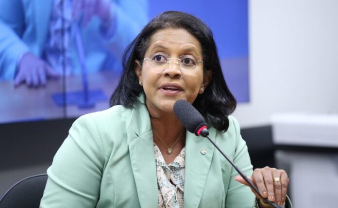 Deputada Gisela Simona fala ao microfone. Ela é uma mulher morena, com cabelo escuro e liso, usa óculo e veste um blazer verde claro