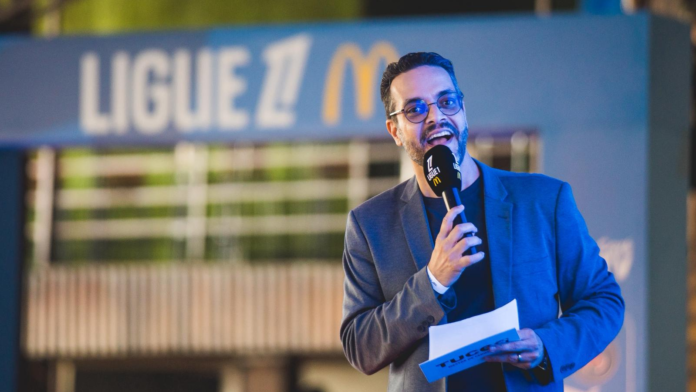 Mestre de cerimônias Tucco apresenta evento da Ligue 1 McDonald’s segurando microfone no palco, com fundo iluminado exibindo o logo da competição.