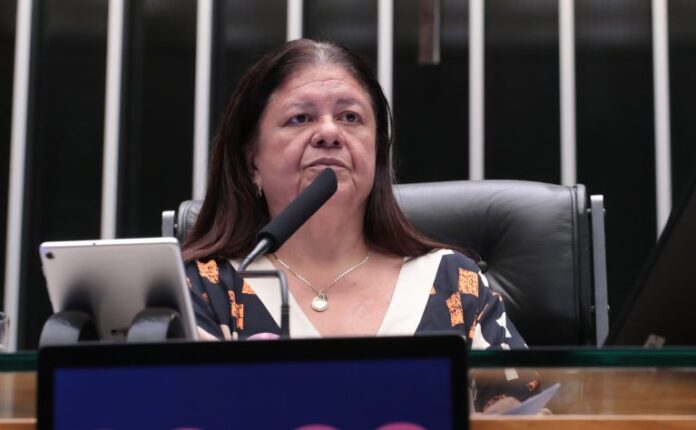 Deputada Laura Carneiro fala ao microfone. Ela é branca, tem o cabelo comprido e escuro, veste uma blusa preta com desenhos em laranja e preto