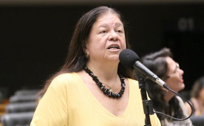 Deputada Laura Carneiro fala ao microfone. Ela é branca, tem o cabelo comprido e escuro, veste uma blusa amarela e colar preto