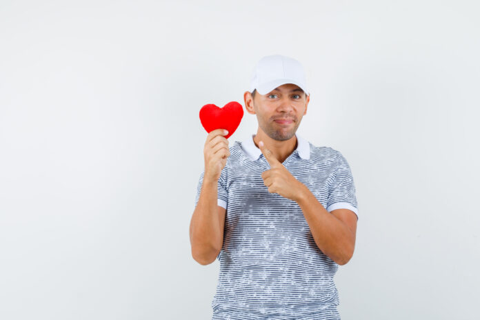Young male pointing at red heart in t-shirt, cap and looking merry. front view. Fonte Freepik