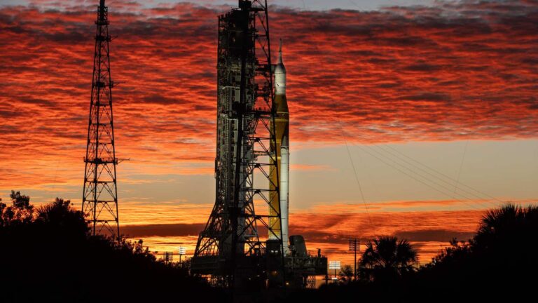 Silhueta do foguete SLS e da nave Orion, da missão Artemis II, na Plataforma de Lançamento 39B do Kennedy Space Center na Flórida. O céu está dramaticamente iluminado com nuvens em tons de vermelho e laranja pelo nascer do sol. Árvores e torres de transmissão aparecem em silhueta no primeiro plano