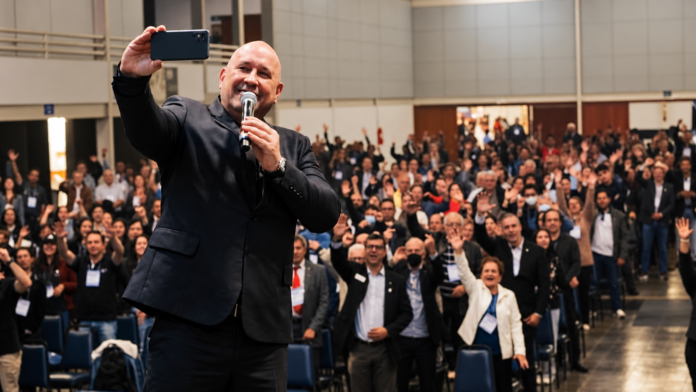 Selfie no palco com plateia entusiasmada Palestrante de terno preto segura um microfone e registra uma selfie diante de uma plateia numerosa em um auditório, com participantes sorrindo e acenando para a câmera.